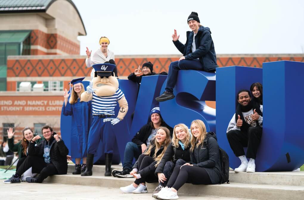Louie the laker and students stand in front, lean against, and sit on top of large blue letters that read 'GVSU'.